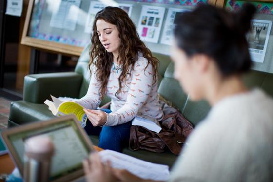 Photo of two female Chatham University students working in the library, one looking at a book and one looking at her computer