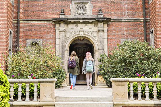 Photo of two female Chatham University students wearing backpacks walking into a red-brick building on Shadyside Campus