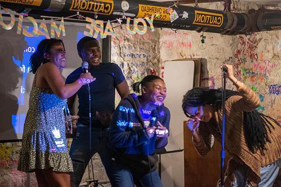 Photo of four Black students laughing and sining on stage with microphones during BIPOC karaoke night. There is a sign behind them that says YOU ARE MAGIC.