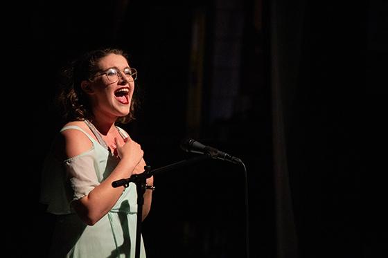 Photo of a Chatham University student standing in a spotlight on a stage singing into a microphone. 